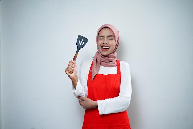 Fröhliche asiatische muslimische Frau in roter Schürze mit Spachtel Kochgeschirr Kochkonzept