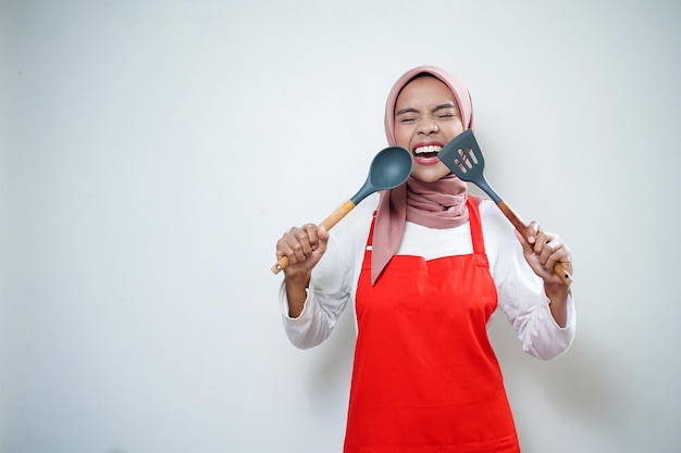 Fröhliche asiatische muslimische Frau in roter Schürze mit Suppenlöffel und Spachtel Kochgeschirr Kochkonzept