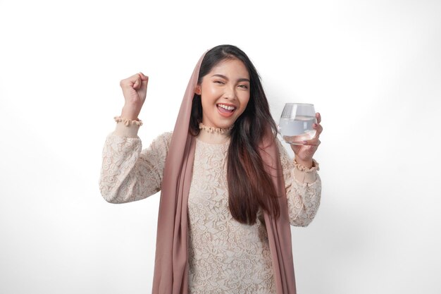 Fröhliche asiatische muslimische Frau mit Kopftuch, Schleier-Hijab, hält ein Glas Wasser mit einem glücklichen, erfolgreichen Gesichtsausdruck, fühlt sich nach dem Fasten erleichtert, isoliert von weißem Hintergrund