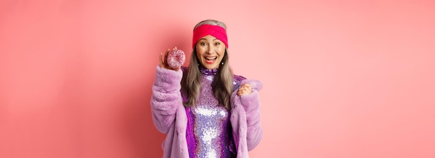 Fröhliche asiatische Seniorin mit grauem Haar, die ein Party-Glitzerkleid trägt, das köstlichen Donut und s zeigt