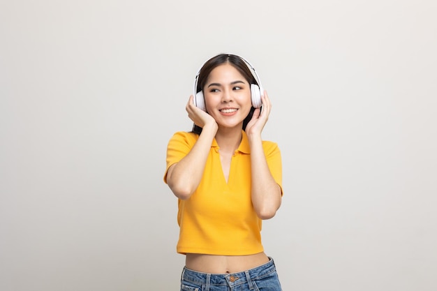 Fröhliche asiatische teenagerin hört musik mit weißem kopfhörer auf isoliert. schöne junge frau im gelben hemd berührt mit der hand einen drahtlosen kopfhörer, der spaß mit der musik im zimmer hat.