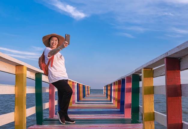 Fröhliche asiatische Touristin, die sich mit dem Smartphone auf der Regenbogen-Holzbrücke am Aussichtspunkt auf das Meer selbst macht