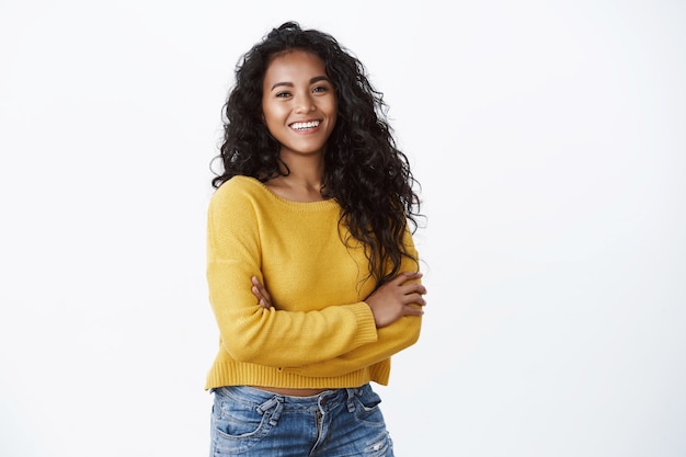 Foto fröhliche attraktive afroamerikanerin hält die arme verschränkt und lacht selbstbewusst, starrt die kamera mit einem entzückenden fröhlichen grinsen an, trägt einen gelben pullover über der weißen wand, wellnesskonzept