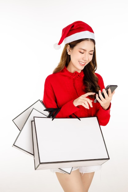 Fröhliche attraktive asiatische Frau, die ein rotes Kleid mit einer Weihnachtsmütze trägt, hält Einkaufstüten auf weißem, isoliertem Hintergrund. Schöne Dame mit langen schwarzen Haaren nutzt Handy-Shopping online.
