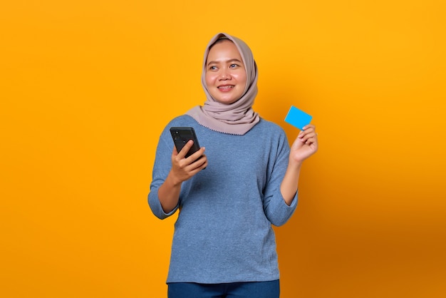 Fröhliche attraktive asiatische Frau, die Handy hält und Kreditkarte über gelbem Hintergrund zeigt