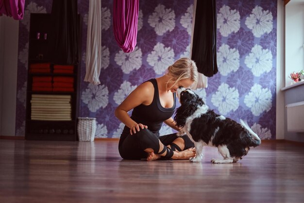 Fröhliche attraktive Frau, die mit ihrem süßen Hund spielt, während sie im Yoga-Fitnessclub auf dem Boden sitzt.