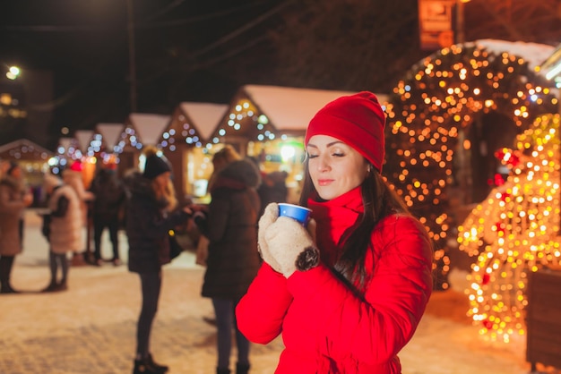 Fröhliche attraktive Frau im roten Wintermantel, die auf dem Weihnachtsmarkt steht, mit geschlossenen Augen träumt und eine Tasse heißes Getränk hält. Lichter des Weihnachtsmarktes mit vielen Menschen im Hintergrund