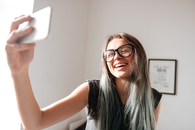 Fröhliche attraktive junge Frau mit Brille, die zu Hause ein Selfie mit dem Handy macht