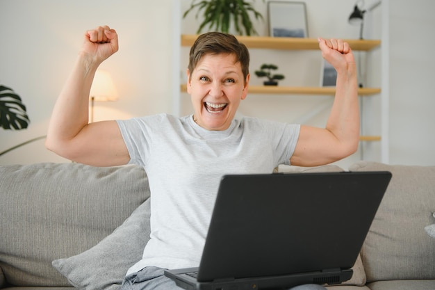 Fröhliche, aufgeregte, grauhaarige, reife Frau, die einen Online-Gewinn mit einem Laptop feiert, der auf den Bildschirm schaut, der zu Hause auf der Couch sitzt, eine Frau mittleren Alters, die sich von unglaublichen guten Nachrichten überrascht fühlt