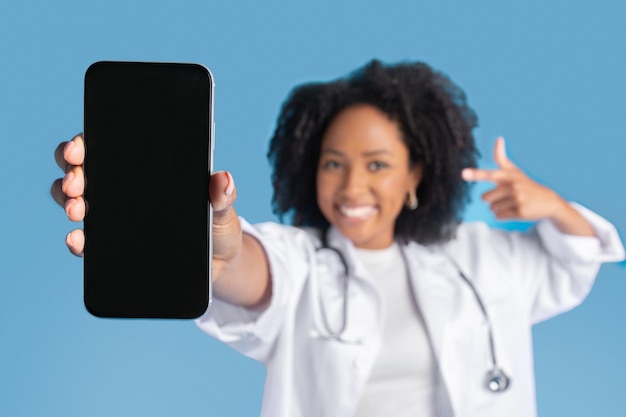 Fröhliche, aufgeregte, junge, lockige Ärztin im Kittel zeigt mit dem Finger auf das Telefon und konzentriert sich auf einen leeren Bildschirm
