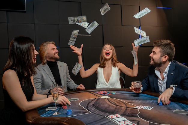 Fröhliche, aufgeregte junge Pokerspielerin, die ein Spiel gewinnt und einen Haufen Papiergeld in die Luft wirft, die am Spieltisch sitzt. Casino, Glücksspielkonzept