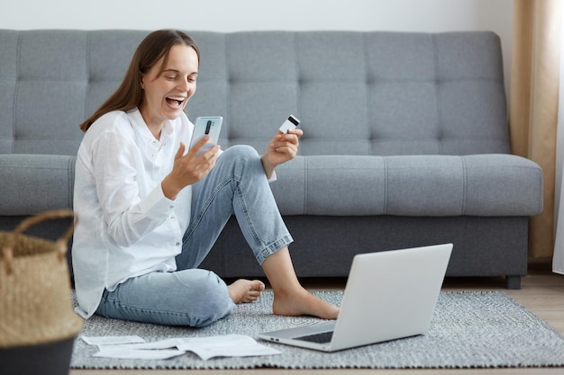 Fröhliche, aufgeregte, zufriedene Frau mit weißem Hemd und Jeans, die mit Laptop und Telefon auf dem Boden in der Nähe des Sofas sitzt und eine Kreditkarte hält, die im Lotto gewinnt und Aufregung ausdrückt