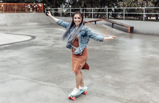 Fröhliche außergewöhnliche tausendjährige Frau mit hellem Make-up und afrikanischen Zöpfen fährt auf einem Penny Board in einem Skatepark