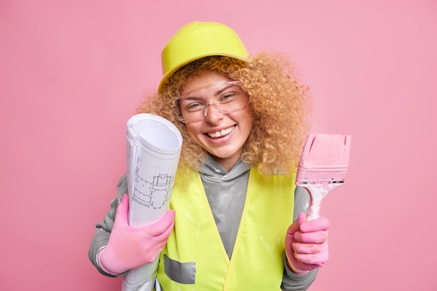 Fröhliche Bauarbeiterin mit guter Laune versucht ihren Bauplan im Leben umzusetzen hält Bauplan und Pinsel zum Streichen von Wänden trägt Schutzhelm und Uniform