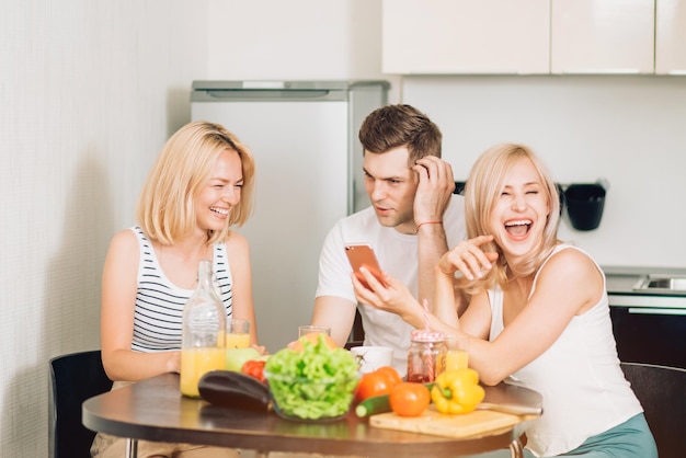 Fröhliche beste Freunde, die lustige Fotos ansehen, Videos in sozialen Netzwerken über das Smartphone ansehen und beim Kochen in der Küche lachen. Drei junge Freunde, die drinnen sitzen und Handy betrachten.