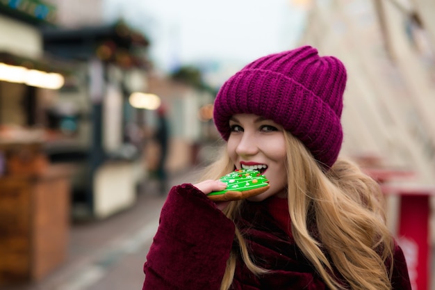 Fröhliche blonde Frau, die Lebkuchenplätzchen isst und auf dem Weihnachtsmarkt in Kiew posiert