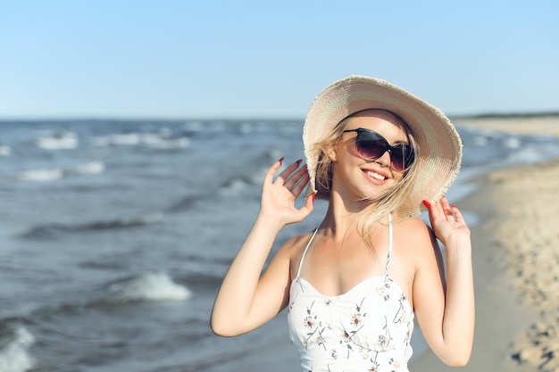 Fröhliche blonde Frau in freier Glückseligkeit am Meeresstrand, stehend mit Sonnenbrille und Hut.