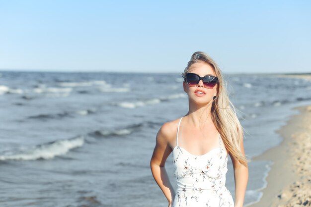 Fröhliche blonde Frau in freier Glückseligkeit am Meeresstrand, stehend mit Sonnenbrille