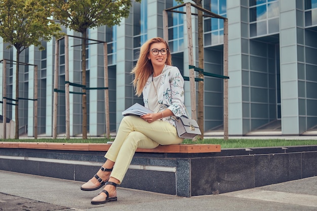 Fröhliche blonde Frau in moderner Kleidung, die mit einem Buch studiert, auf einer Bank im Park gegen einen Wolkenkratzer sitzt.