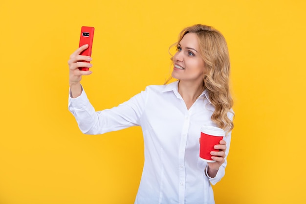 Fröhliche blonde Frau mit Kaffeetasse macht Selfie am Telefon auf gelbem Hintergrund