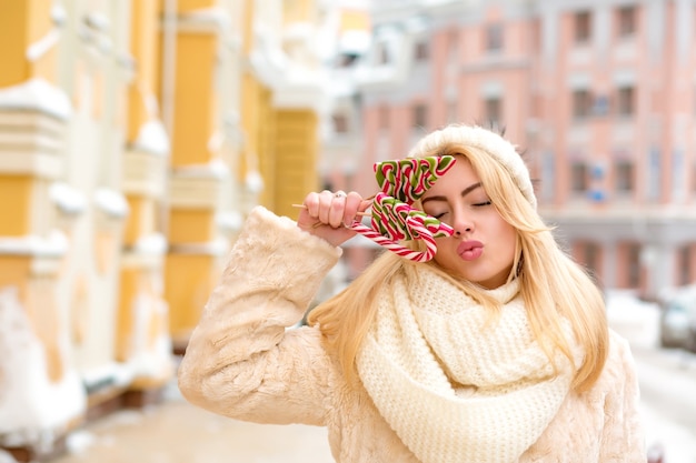 Fröhliche blonde Frau mit langen Haaren, die eine warme Strickmütze trägt und köstliche Weihnachtssüßigkeiten hält