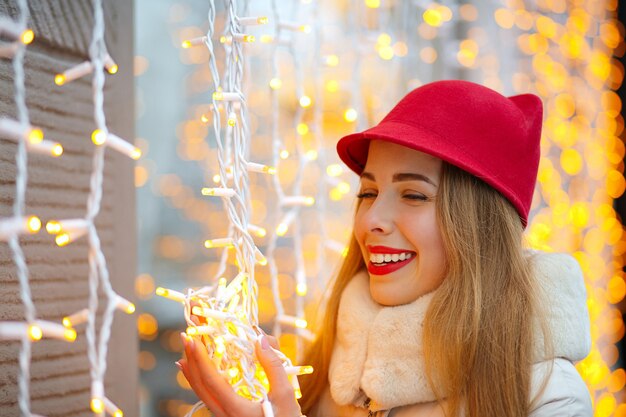 Fröhliche blonde Frau mit rotem Hut posiert auf der Straße mit leuchtenden Girlanden. Platz für Text