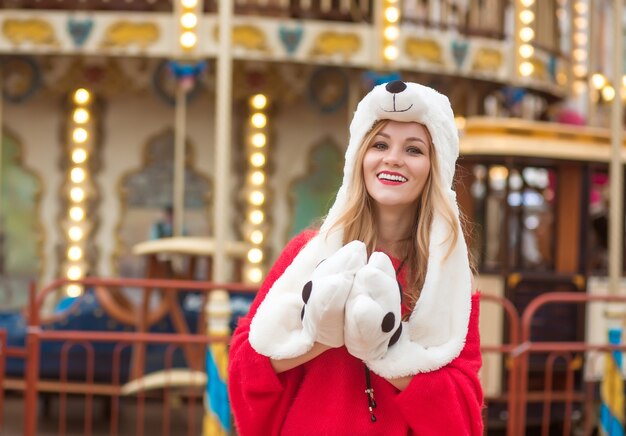 Fröhliche blonde frau mit rotem strickpullover und lustigem hut, die im hintergrund des karussells mit lichtern posiert