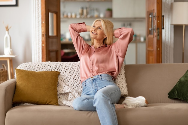 Fröhliche blonde reife Frau sitzt auf der Couch und entspannt sich zu Hause