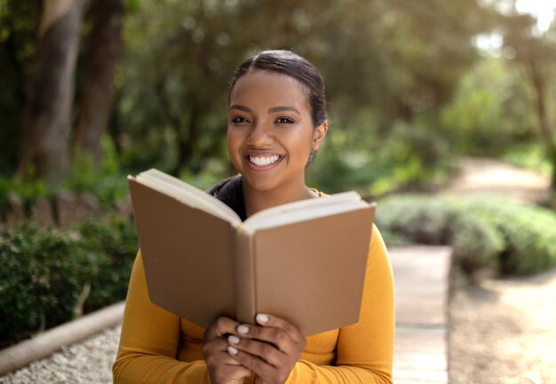 Fröhliche brasilianische Studentin, die im Park im Freien sitzt und ein Buch liest, das in die Kamera schaut und lächelt