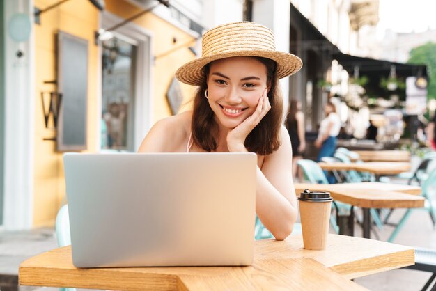 fröhliche brünette Frau, die lächelt und Laptop benutzt, während sie im Straßensommercafé mit Kaffee zum Mitnehmen sitzt?