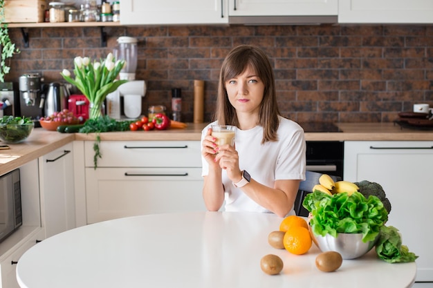 Fröhliche brünette Frau, die zu Hause mit hausgemachtem Glas-Smoothie und gesunden Früchten sitzt Vegane Mahlzeit und Detox-Konzept Mädchen mit weißem T-Shirt, das frischen Cocktail trinkt