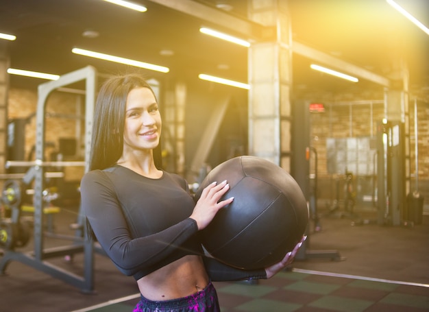 Fröhliche brünette Frau in der Sportbekleidung, die Medizinball in ihrer Hand hält und an der Kamera aufwirft.