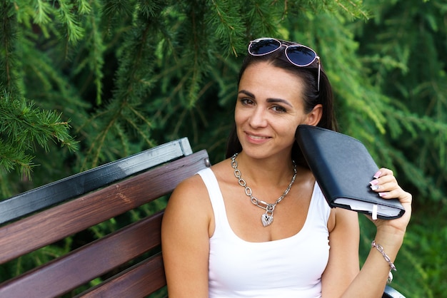 Fröhliche brünette Frau in Freizeitkleidung und Sonnenbrille sitzt auf einer Holzbank im Park gegen ...