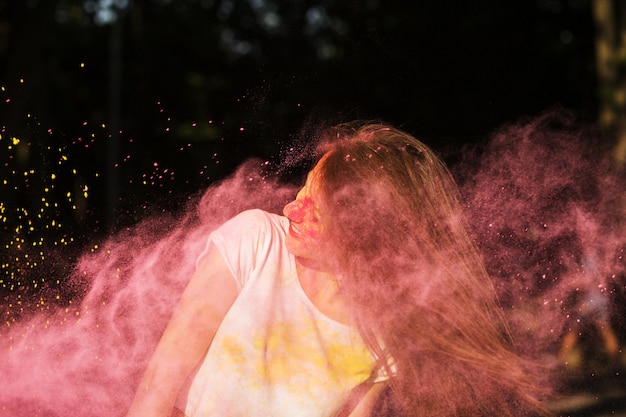 Fröhliche brünette Frau mit Haaren im Wind posiert mit rosa Pulver Holi, das um sie herum explodiert