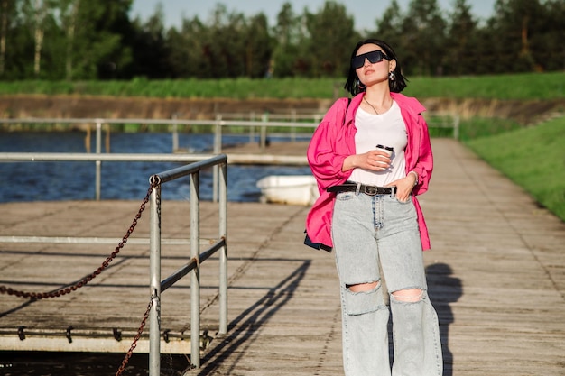 Fröhliche brünette Frau mit Sonnenbrille, die an sonnigen Tagen in der Nähe des Sees auf einem Holzsteg spazieren geht