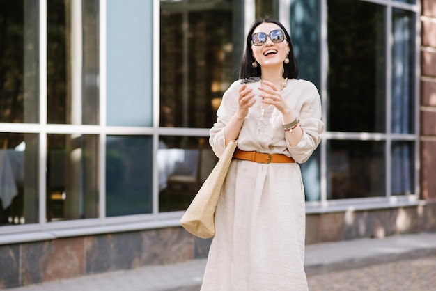 Fröhliche brünette junge Frau mit Sonnenbrille und Tasche mit Kaffee, die in der Stadt spazieren geht. Lifestyle-Porträt einer lächelnden Frau