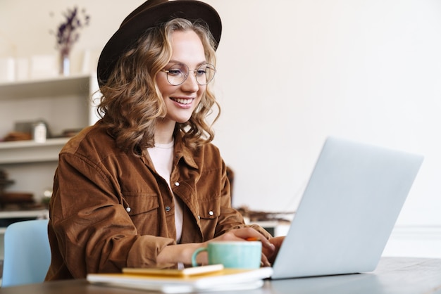 Foto fröhliche charmante frau mit brille und hut, die mit laptop arbeitet, während sie am tisch sitzt