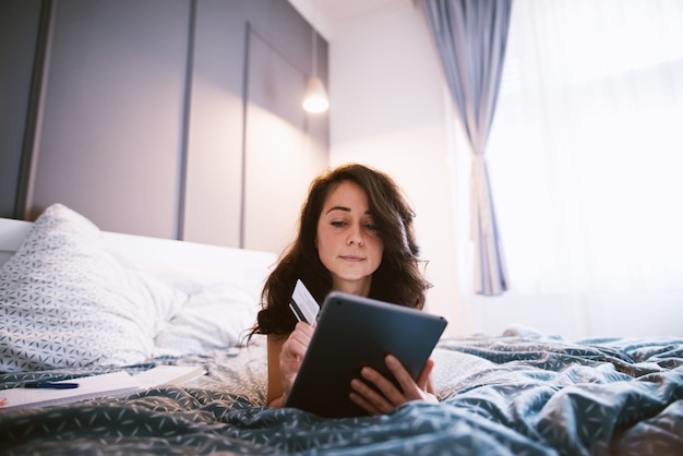 Fröhliche charmante Frau mittleren Alters, die Bankkarte hält und online auf einer Tablette kauft, während sie auf dem Bett liegt.