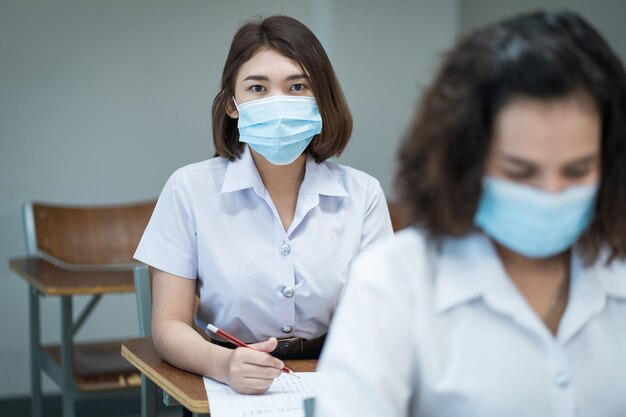 Fröhliche College-Studenten im Klassenzimmer tragen schützende Gesichtsmasken und verwenden Antiseptika zur Vorbeugung von Coronaviren während der Coronavirus-Pandemie. Gruppe von Schülern, die im Unterricht Schutzmasken tragen.