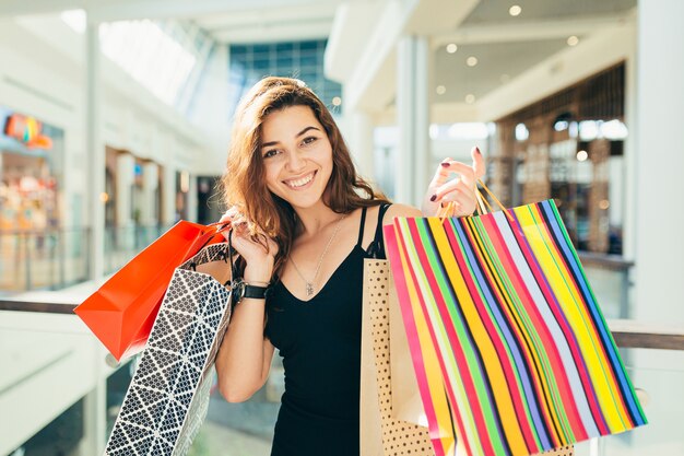 Fröhliche Dame im stilvollen schwarzen Kleid, das bunte Einkaufstaschen beim Gehen am Laden weiterführt