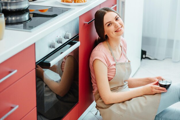Foto fröhliche dame in einer schürze lächelt, während sie in der küche am herd sitzt und eine tasse in den händen hält. website-banner