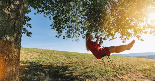 Fröhliche Dame in rotem Sommerkleid, die sich in ihrer Schaukel in einer sonnigen, goldenen Landschaft zurücklehnt