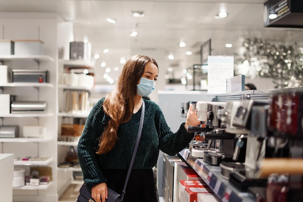 Fröhliche Dame mit Gesichtsmaske wählt während der Covid-Pandemie eine Kaffeemaschine im Einkaufszentrum