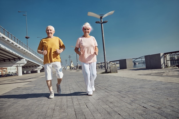 fröhliche Dame und ihr glücklicher Ehemann, die zusammen den Kai entlang joggen