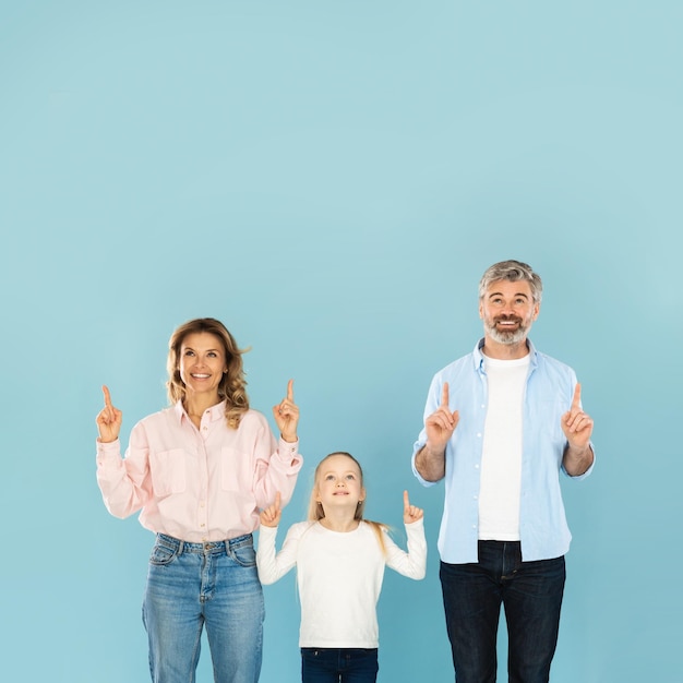 Fröhliche dreiköpfige Familie, die mit dem Finger nach oben auf blauen Hintergrund zeigt