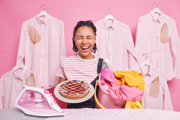 Fröhliche dunkelhäutige afroamerikanerin, die an der hausarbeit beteiligt ist, hat viele verantwortungen hält einen eimer mit wäsche köstlich gebackener kuchen posiert in der nähe des bügelbretts drückt positive emotionen