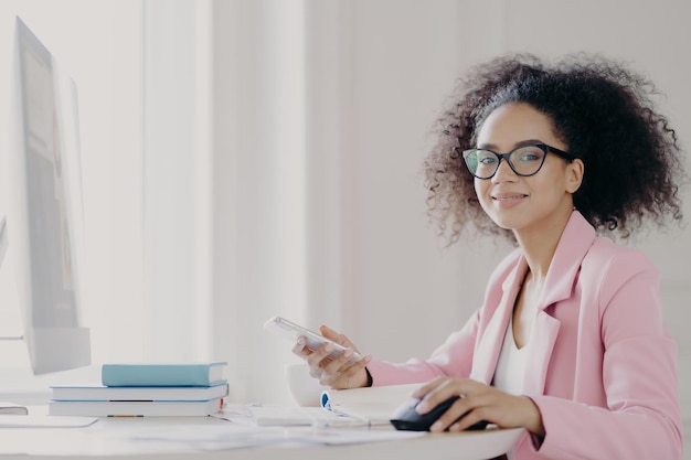 Fröhliche dunkelhäutige Dame hält Handy in der Hand wartet auf Anruf sitzt vor Computer trägt optische Brille und eleganten Anzug posiert in ihrem Kabinett und ist bei der Arbeit beschäftigt Tech-Beruf