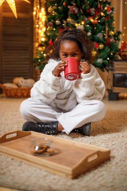 Fröhliche dunkelhäutige Teenagerin hält einen roten Becher mit Kakao in den Händen in der Nähe des Weihnachtsbaums zu Hause