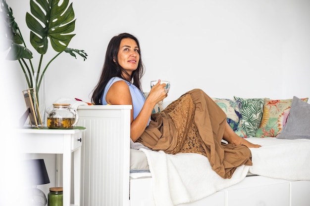 Fröhliche, entspannte, elegante brünette Frau, die auf einer bequemen Couch Tasse Tee sitzt und träumt, Zeit allein zu verbringen?