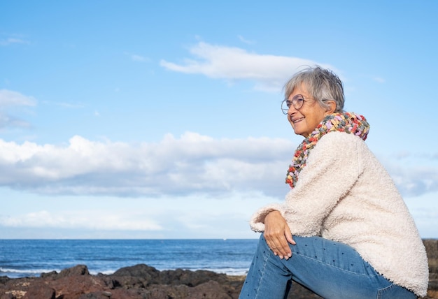 Fröhliche, entspannte, erwachsene Seniorin, die am Strand sitzt und einen Pullover trägt, der wegschaut